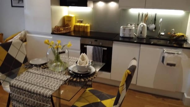 Een kleine keuken met een magnetron, broodrooster en gootsteen. Er staat een eettafel met een tafelkleed met een patroon en een vaas met gele bloemen.