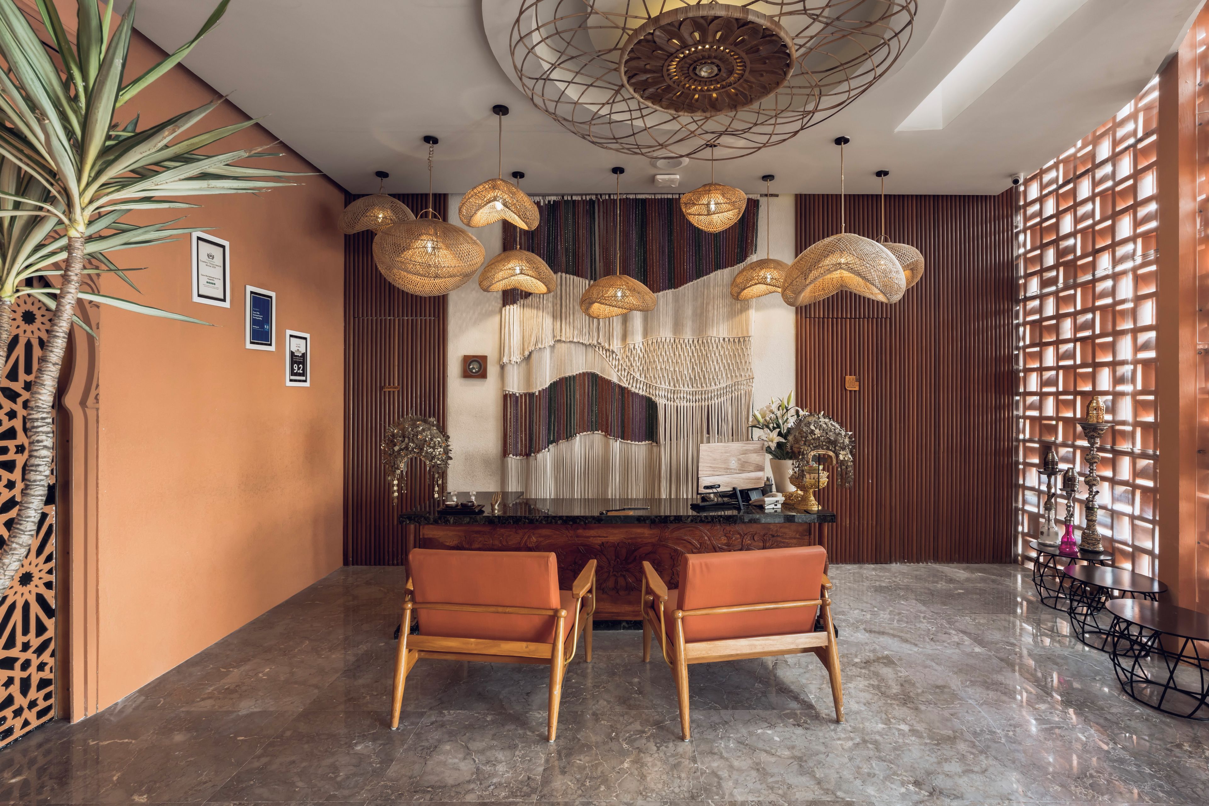 Zona de recepción con mostrador de madera, dos sillones naranja, una pared decorativa con listones verticales y lámparas colgantes tejidas.