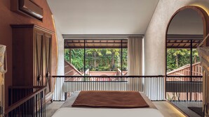 A bedroom with a large bed, a view of a balcony with a wooden railing, and a window overlooking greenery.