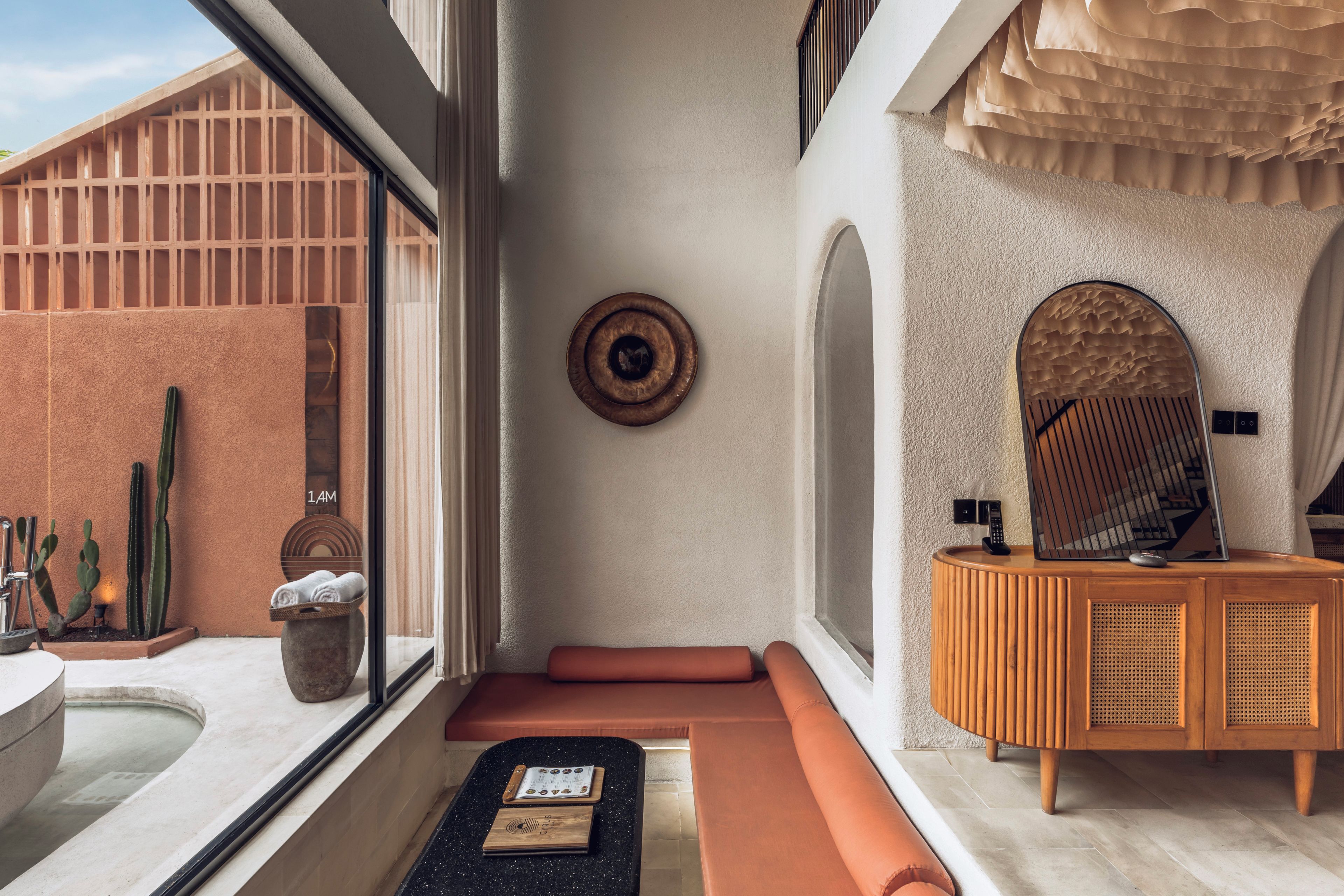 A modern bathroom with a bathtub, a wooden vanity, a mirror, and a window with a view of cacti.