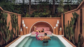 Una piscina con una persona y una mujer en una cama, rodeada de cactus y un muro de ladrillo.