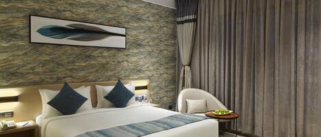 A modern hotel room with a large bed, a headboard with a light fixture, a bedside table with a phone and a pen, and a small table with a plate of apples.