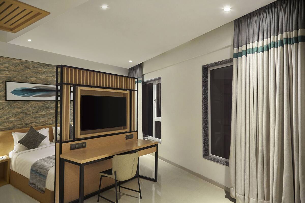 A modern hotel room with a bed, a desk with a chair, a television, and a large window with curtains.