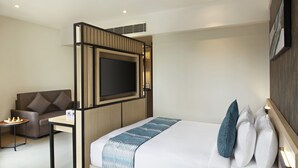 Una habitación de hotel moderna con una cama, un televisor montado en una separador de madera, un sofá, una mesita con una bandeja de fruta y una mesita de noche con una lámpara.