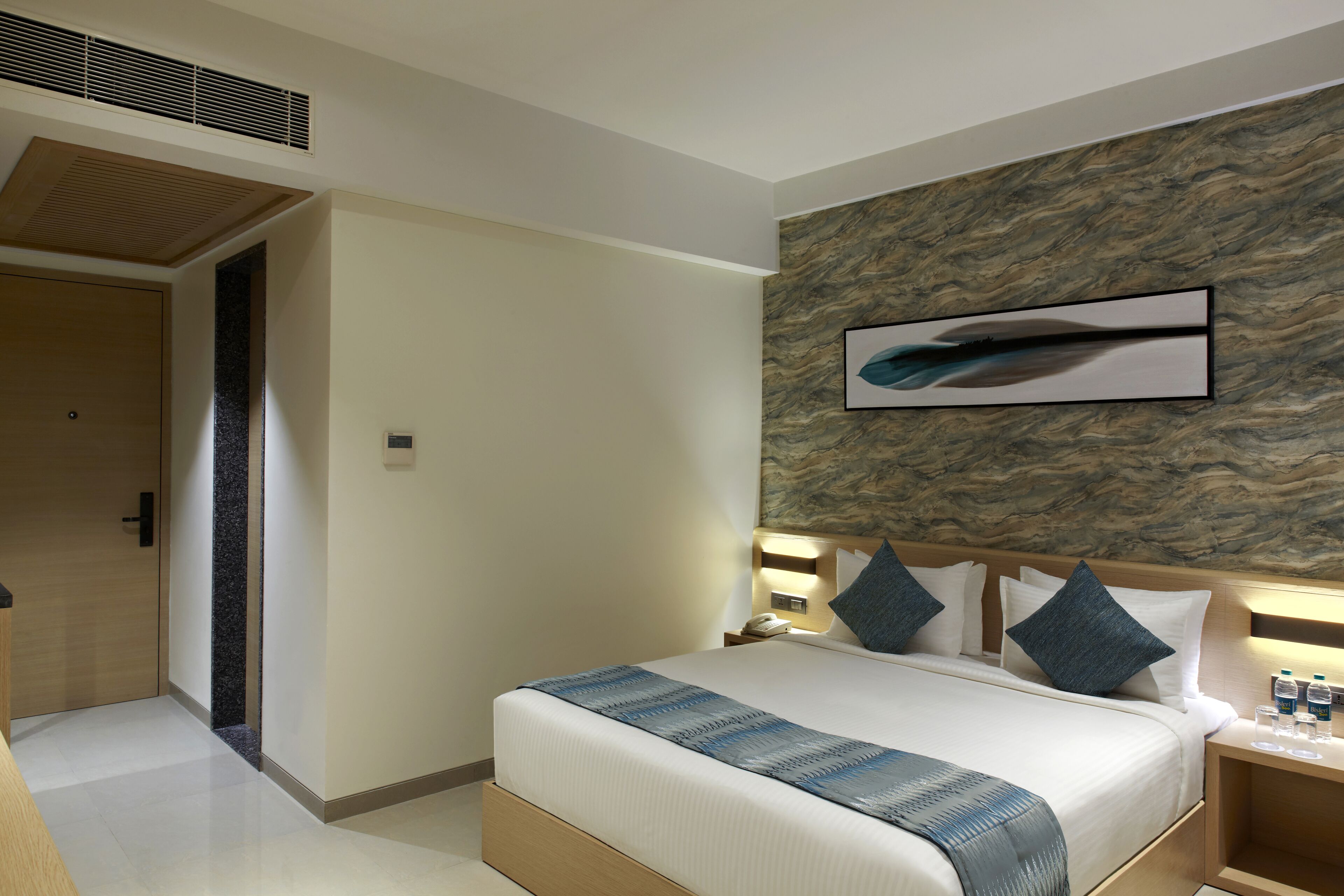 A modern hotel room with a large bed, a headboard with a built-in light, a wall-mounted artwork, and a wooden door.