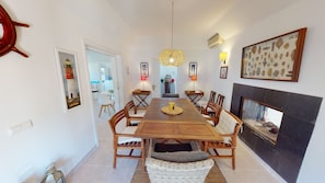 Uma sala de jantar com mesa de madeira, cadeiras, luminária pendente no centro e lareira.