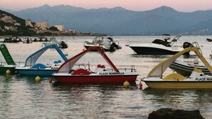 Un puerto deportivo con barcos amarrados, un promontorio rocoso en el agua y montañas al fondo.