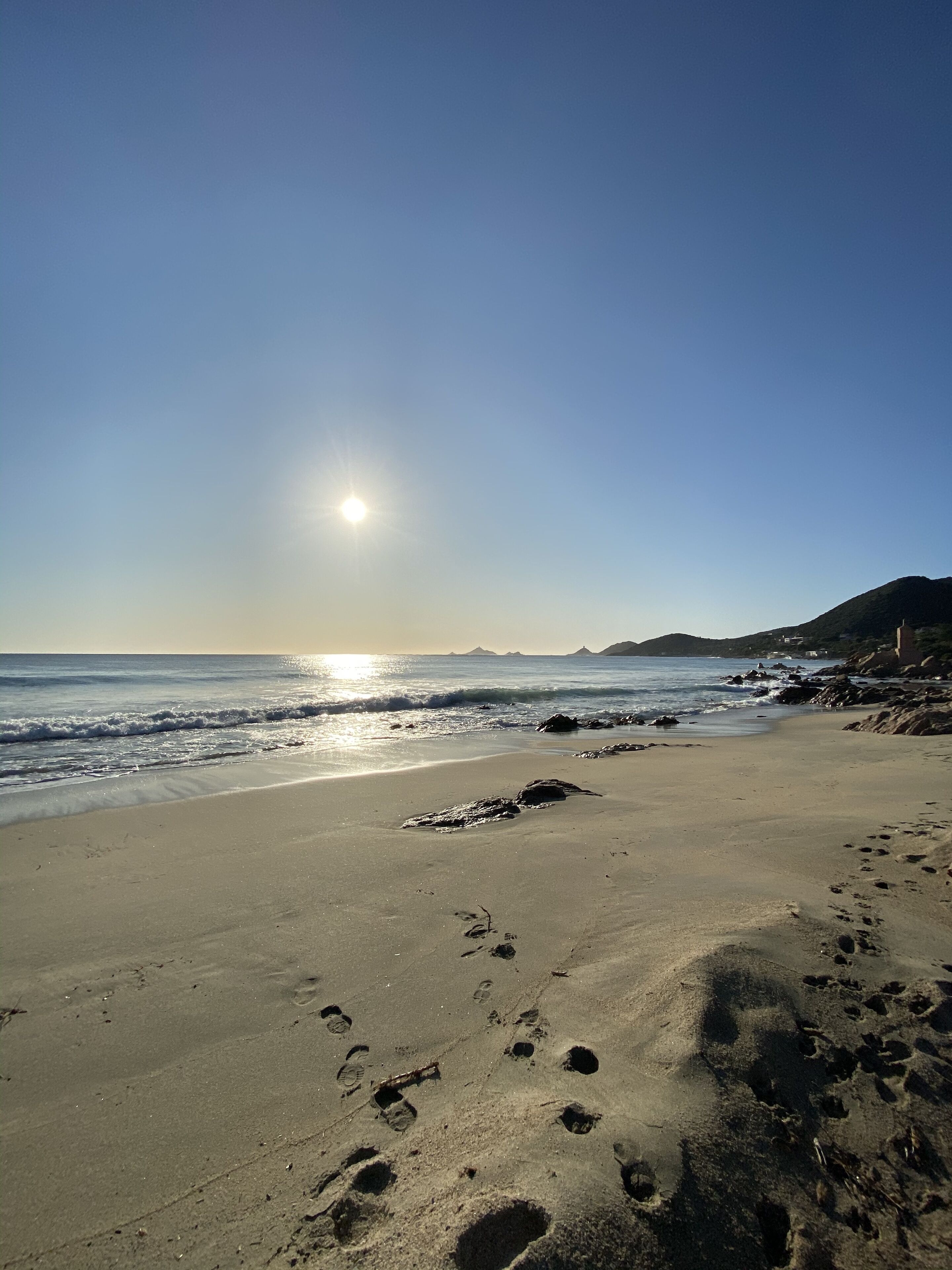 A beach with footprints and waves, a clear sky, and distant mountains.