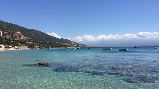 A coastal scene with clear turquoise water, boats, and buildings on a hillside. - Villa with swimming pool and directly by the sea on the Rte des Sanguinaires (Ajaccio)