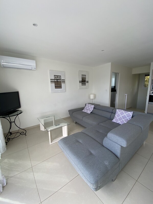 A modern living room with a grey sectional sofa, a glass coffee table, a flat-screen TV, and two wall-mounted air conditioning units.