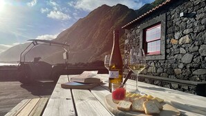 A wooden table with a bottle of white wine, a glass, and bread slices. A stone building with a window is in the background. - Stylish 1 Bedroom Stone House On Ocean Front - São Jorge, Azores (Bela Lua) (Calheta)