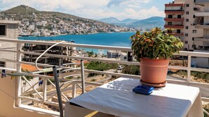 Un balcón con planta en maceta, una mesa con un objeto azul, y vista a una ciudad costera con edificios y mar azul cristalino.
