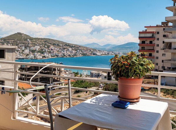 A balcony with a potted plant, a table with a blue object, and a view of a coastal cityscape with buildings and a clear blue sea. - Mariana Apartments (Sarandë)