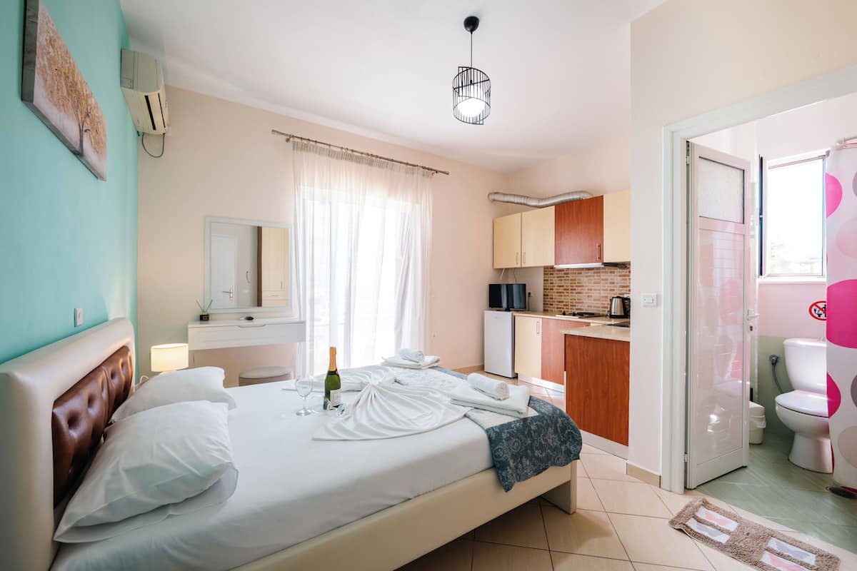 A hotel room with a bed, a bedside table, a window with curtains, a bathroom with a toilet and sink, and a kitchenette area.
