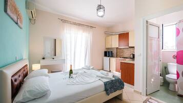 A hotel room with a bed, a bedside table, a window with curtains, a bathroom with a toilet and sink, and a kitchenette area.