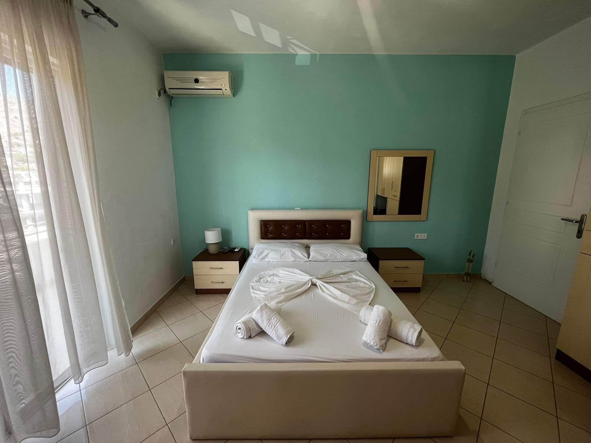A hotel room with a bed, two bedside tables, an air conditioning unit, a mirror, and a door.