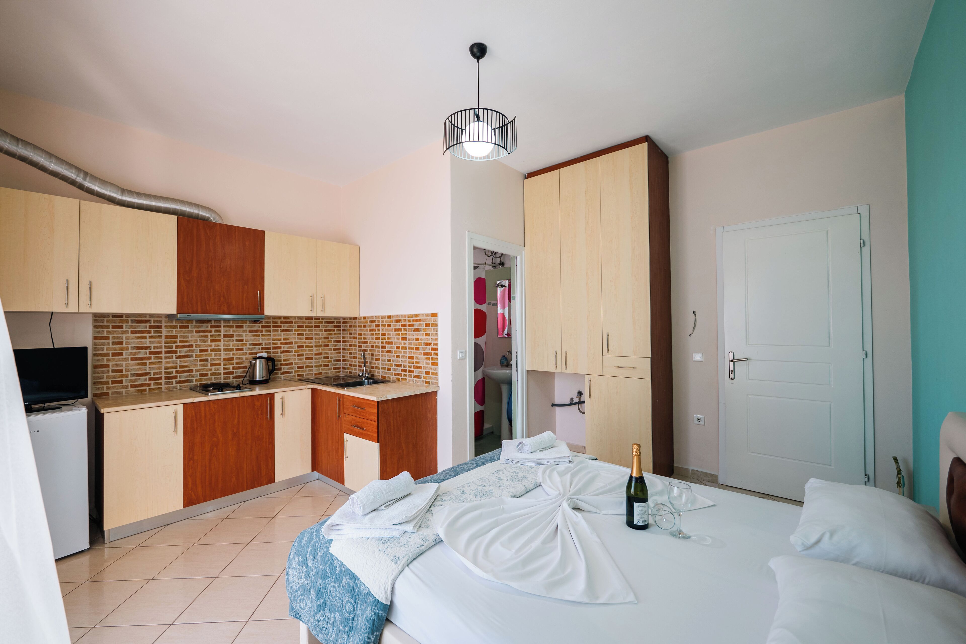 A compact hotel room with a kitchenette, a bed with towels and a bottle of champagne, and a door to the bathroom.