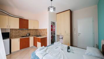 A compact hotel room with a kitchenette, a bed with towels and a bottle of champagne, and a door to the bathroom.