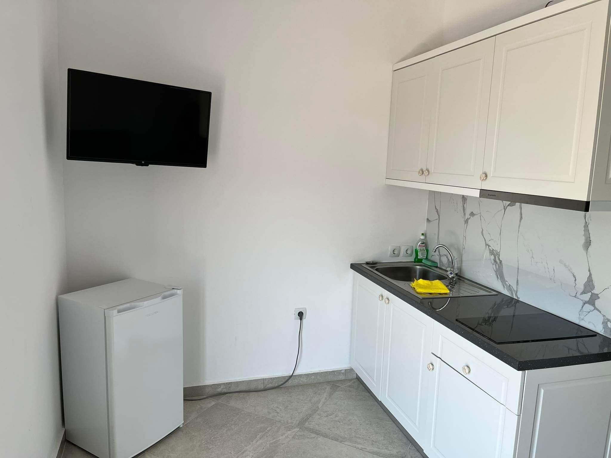 Una cocina compacta con armarios blancos, encimera negra, microondas integrado y televisor montado en la pared.
