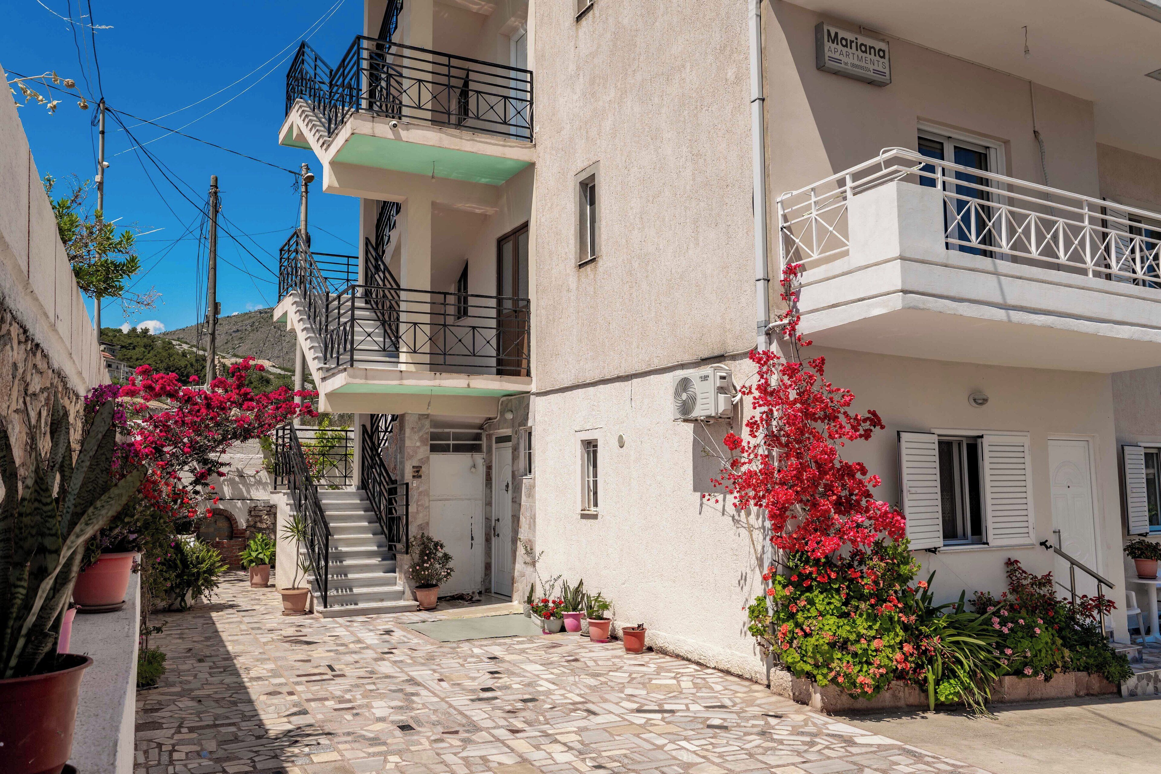 Un immeuble sur lequel on peut lire l’inscription « Mariana Apartments ». Un escalier mène à un balcon orné d’une rambarde en métal. Des plantes et des fleurs en pot sont disposées autour de l’immeuble.
