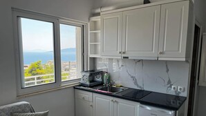 Una cocina compacta con armarios blancos, encimera negra y vista al mar por una ventana.