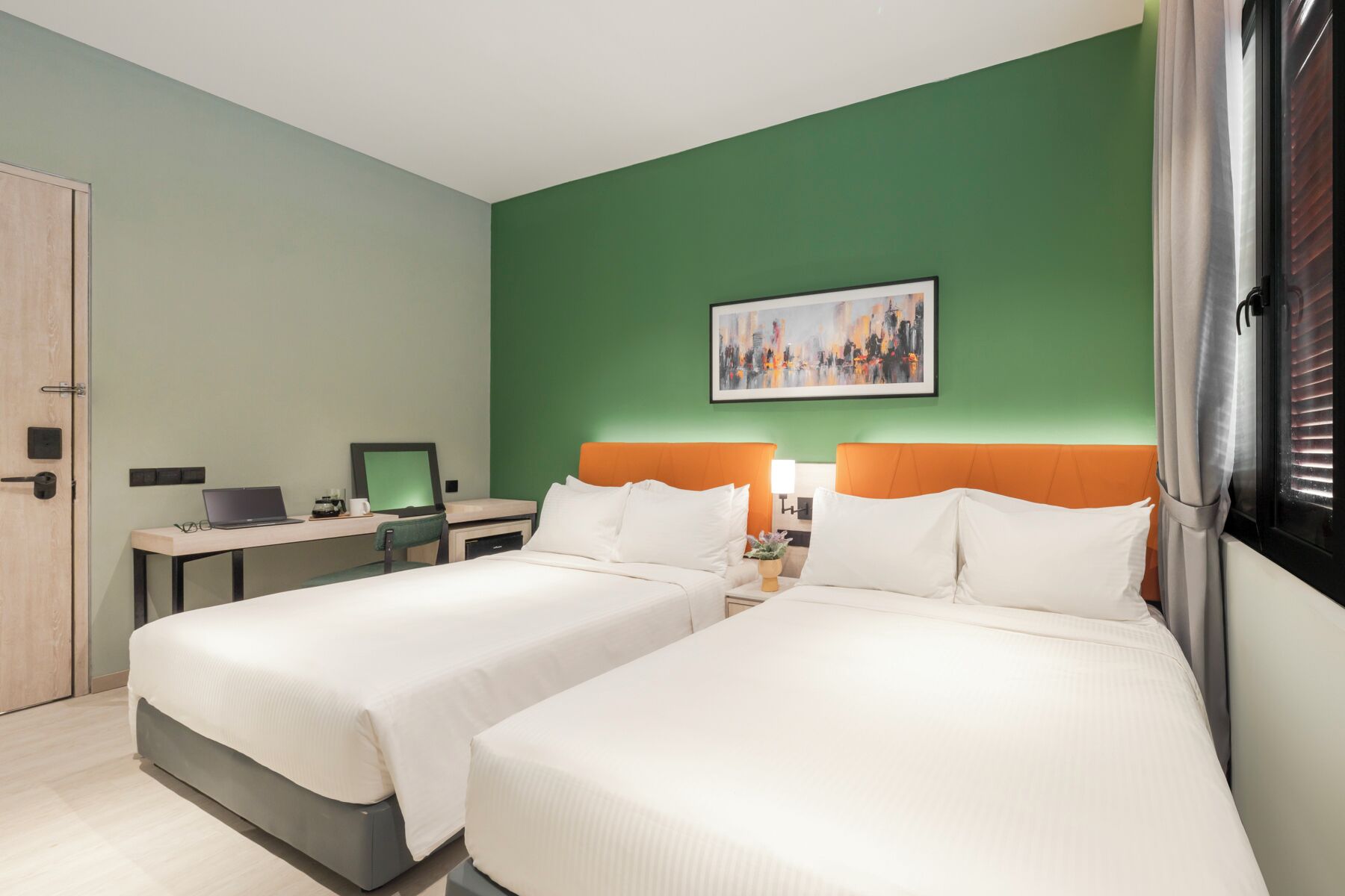 A hotel room with two beds, a desk with a laptop, a painting on the wall, and a window with blinds.