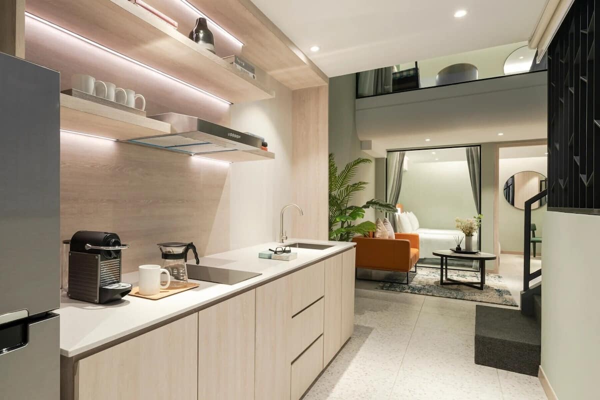 A modern kitchen with a built-in oven, a coffee machine, and a sink. There is a seating area with a sofa and a small table.
