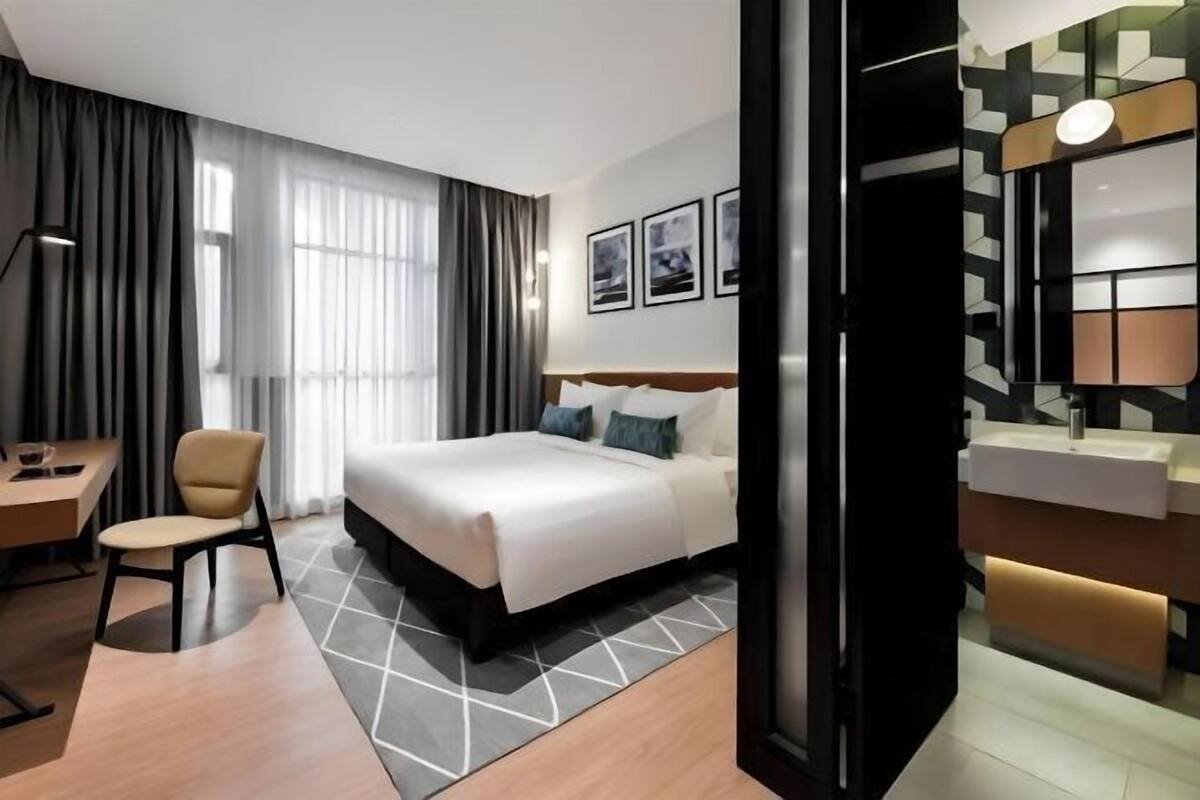 A hotel room with a bed, a desk, a chair, a large window with curtains, and a wall-mounted light fixture.