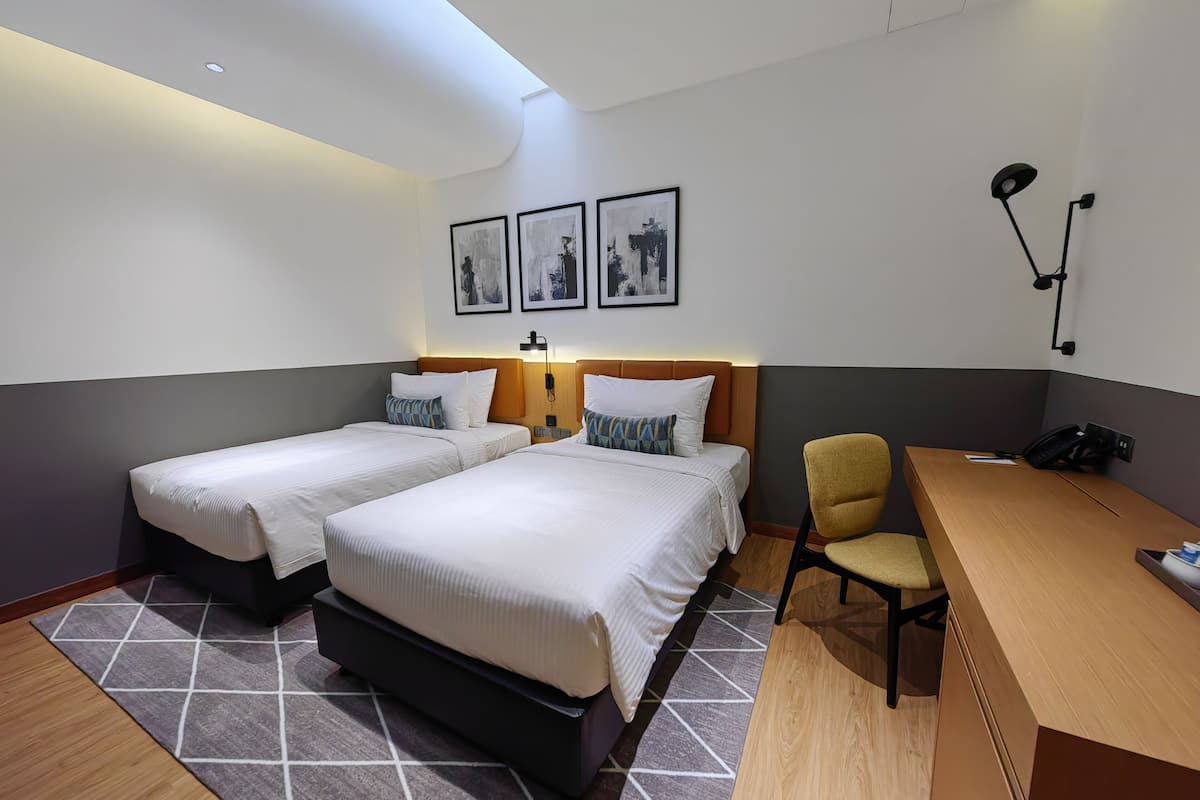 A hotel room with two beds, a desk, a chair, and a desk lamp. There are framed pictures on the wall and a phone on the desk.