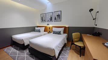 A hotel room with two beds, a desk, a chair, and a desk lamp. There are framed pictures on the wall and a phone on the desk.