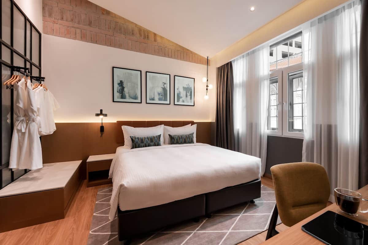 A modern hotel room with a large bed, a desk with a chair, a wardrobe, and a window with curtains.