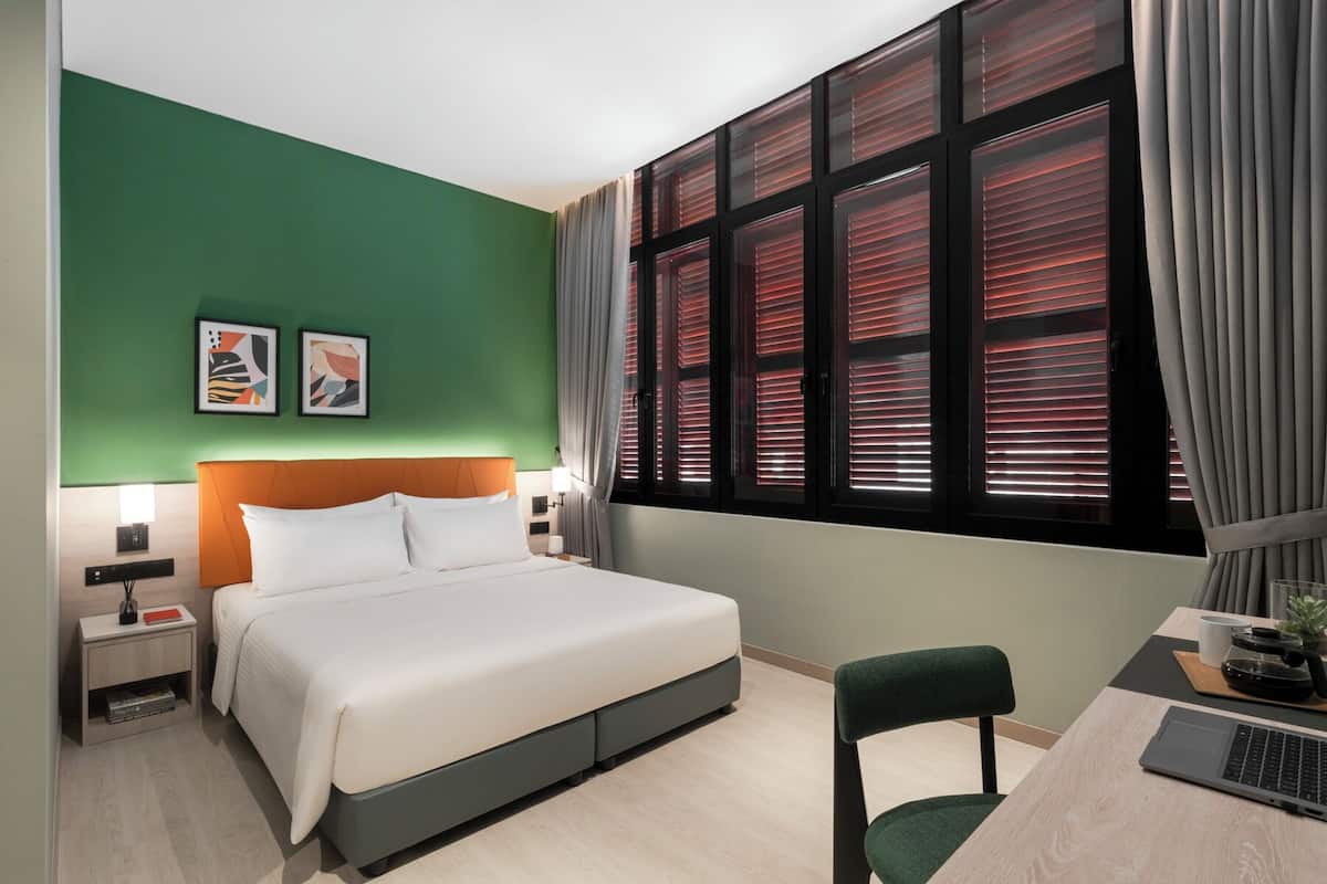 A hotel room with a bed, a desk with a laptop, a chair, two framed artworks on the wall, and a window with blinds.