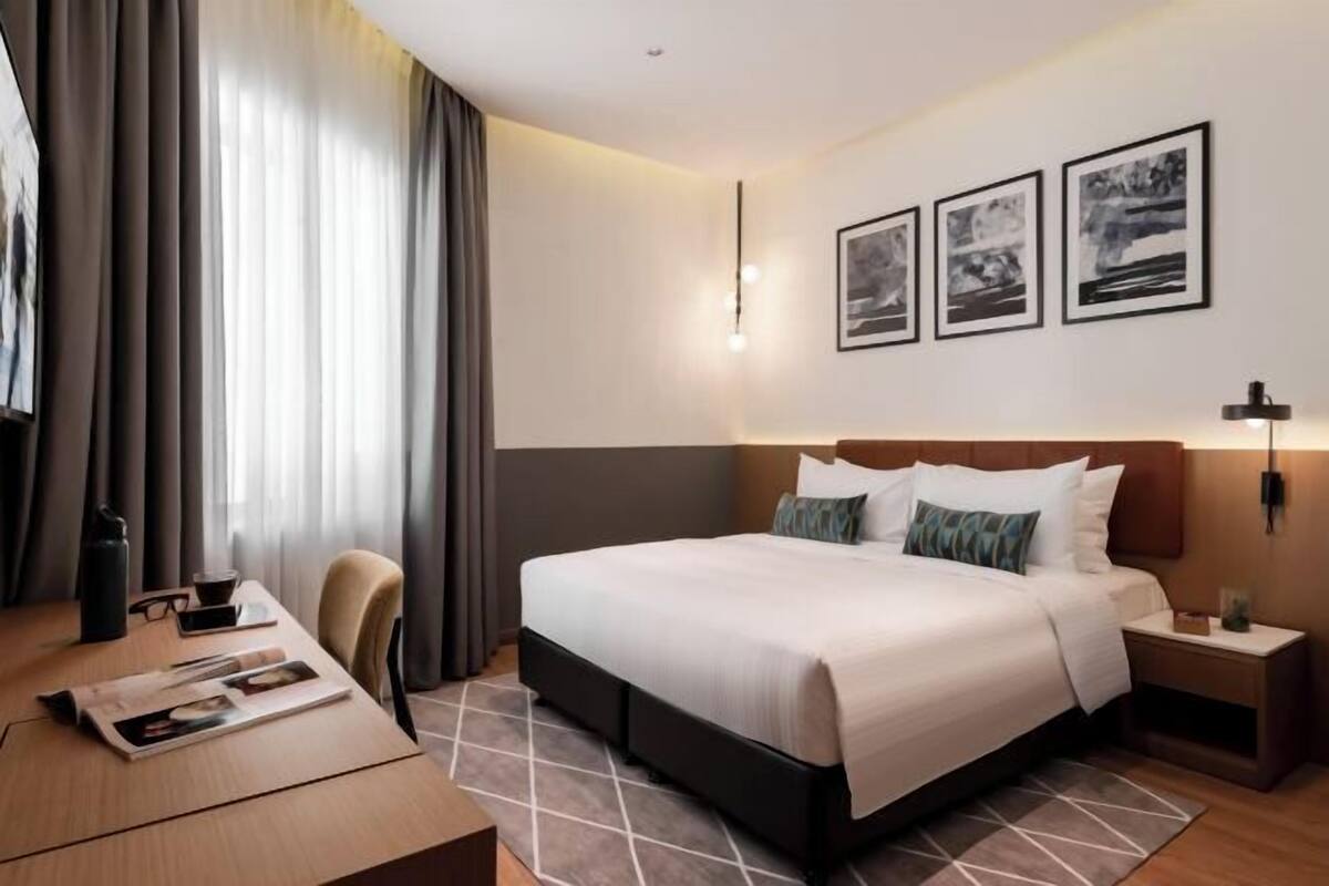 A hotel room with a bed, bedside table, lamp, and a desk with a chair.