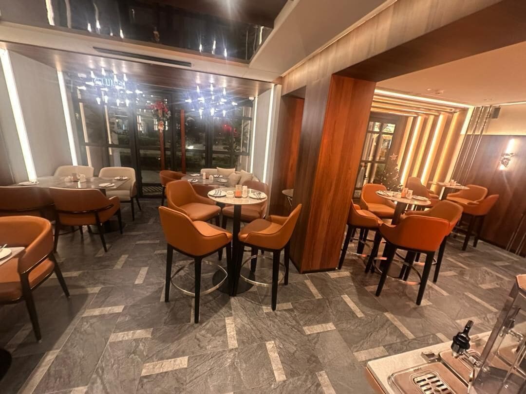 A restaurant with round tables and orange chairs, a herringbone floor pattern, and a view through large windows.