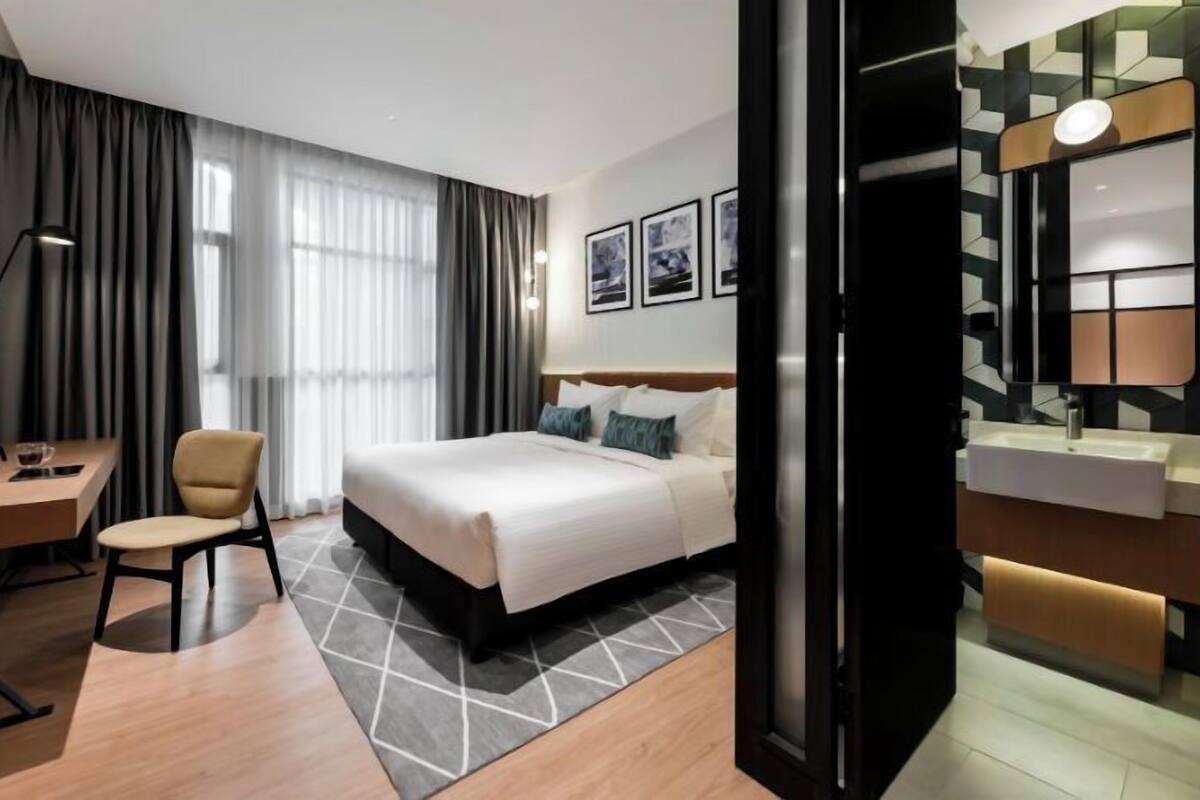 A modern hotel room with a large bed, a desk with a chair, a vanity with a mirror, and a geometric-patterned rug.