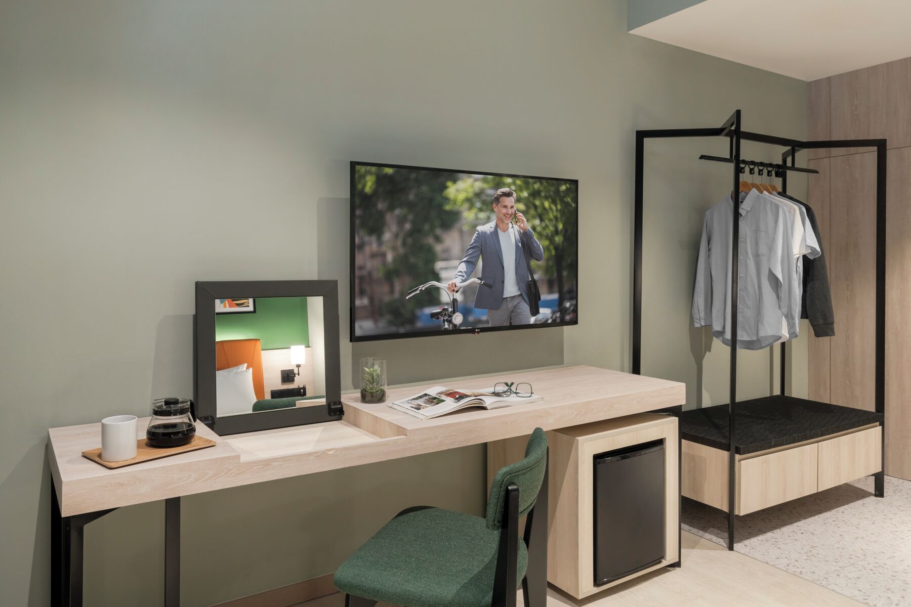 A modern hotel room with a large TV, a desk with a coffee maker, a chair, and a clothing rack.