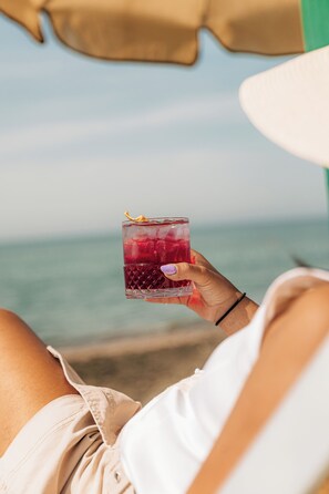 A person holding a drink with a straw, wearing a hat, and sitting under an umbrella.