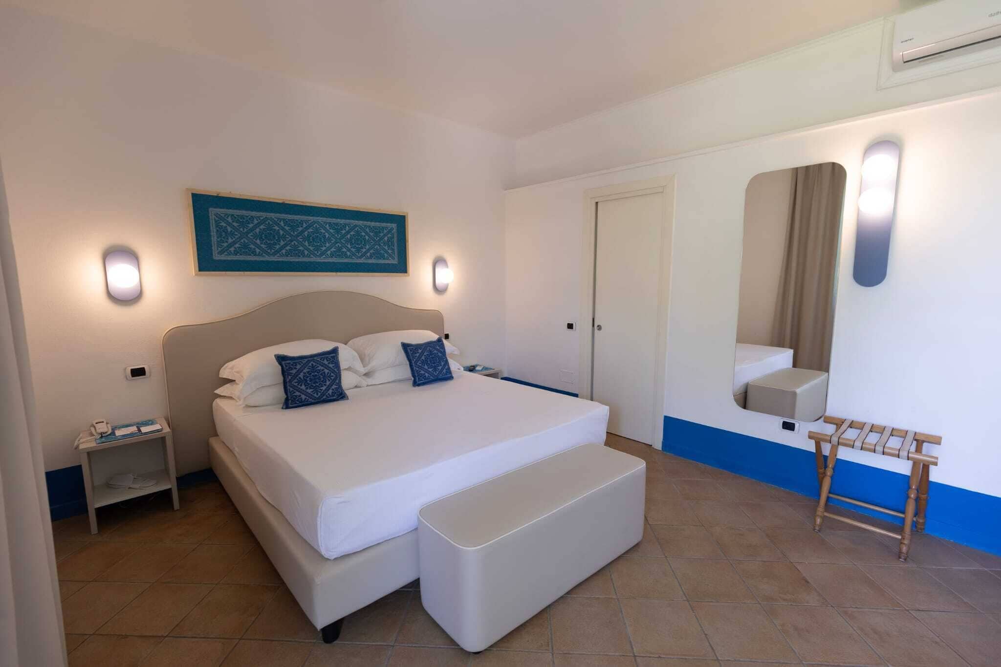 Un dormitorio con una cama grande, mesitas de noche, un banco, un espejo y una puerta.