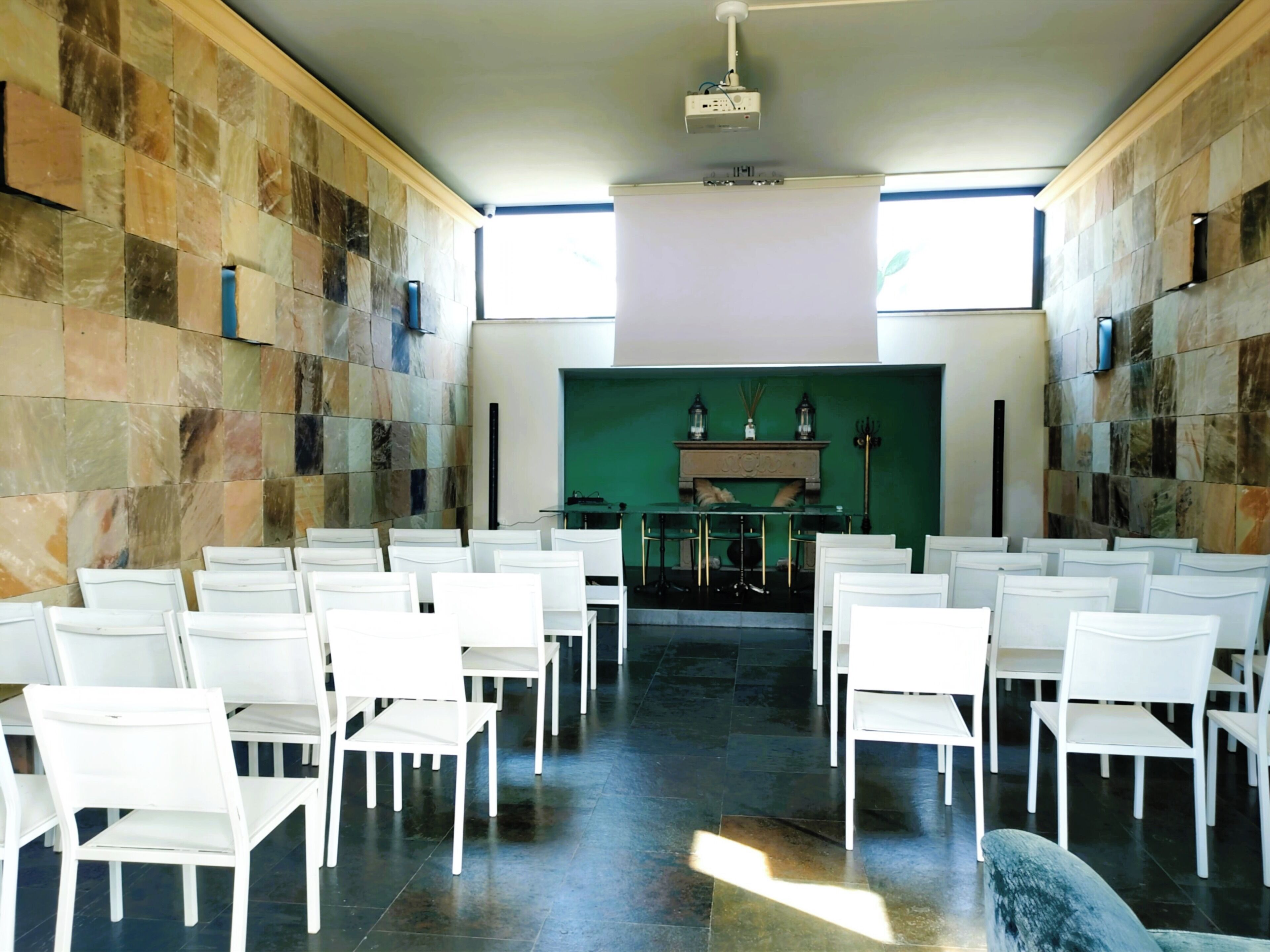A conference room with white chairs arranged in rows, a green screen at the front, and a projector mounted on the ceiling.