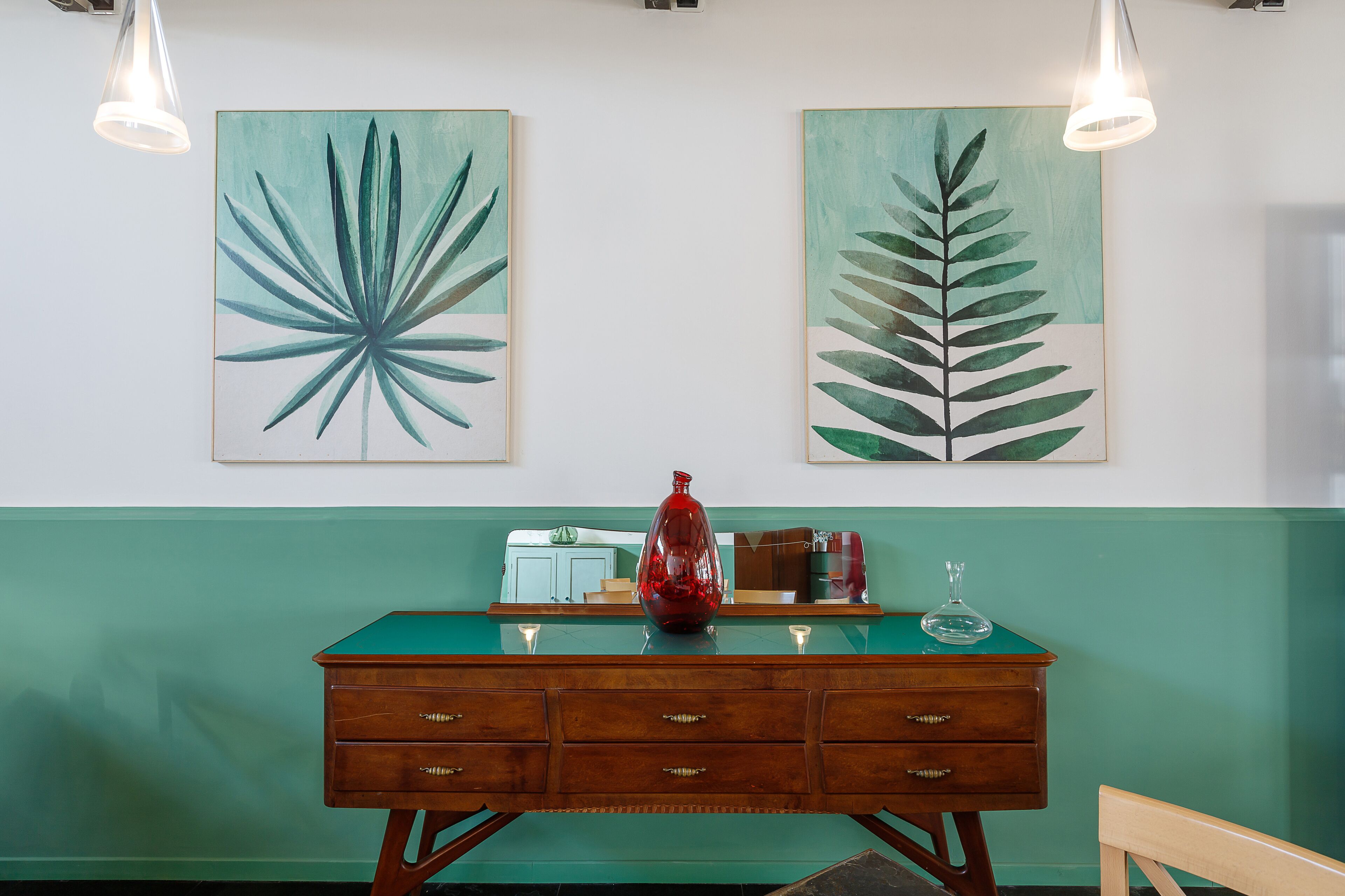 A room with a green wall, a wooden dresser with drawers, a glass-top table, and two wall-mounted paintings of leaves.