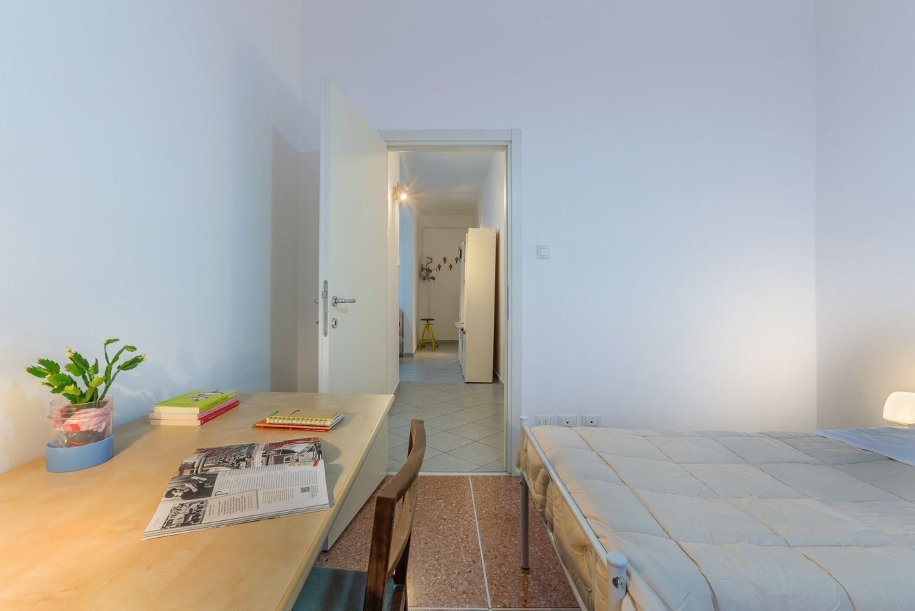 A small, tidy room with a wooden desk, a bed, and a door leading to another room.