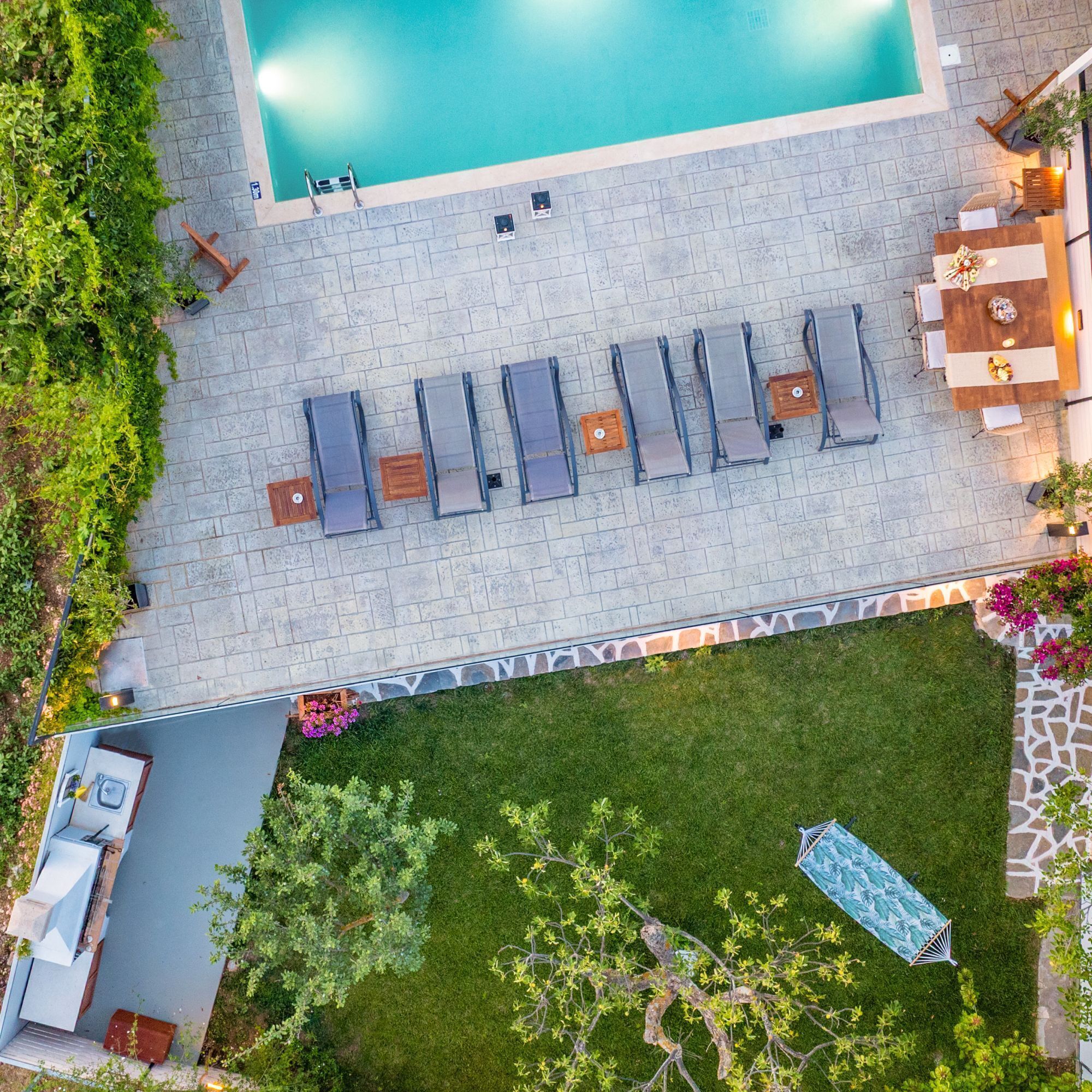 An aerial view of a pool area with a row of sun loungers, a small pool, and a grassy lawn with a hammock.