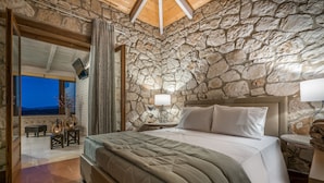 Un dormitorio con pared de piedra, una cama grande, una mesita de noche, una lámpara y vistas al exterior.