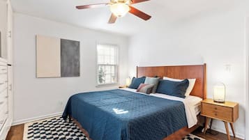 Ein Schlafzimmer mit Bett, Nachttisch, Deckenventilator, Fenster und einem Wandbild.