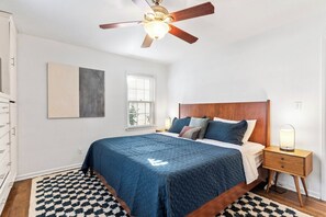 A bedroom with a bed, a nightstand, a ceiling fan, a window, and a wall-mounted artwork.