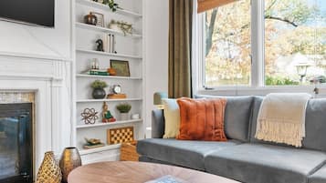 Ein Wohnzimmer mit grauem Sofa, Kamin, Bücherregal und einem Fenster mit Blick auf Bäume.