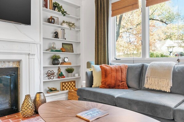 A living room with a grey sofa, a fireplace, a bookshelf, and a window with a view of trees.
