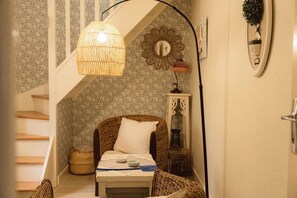 Une pièce chaleureuse comprenant un lit, un fauteuil en osier, une petite table, un luminaire suspendu, un miroir décoratif et une plante fixée au mur.