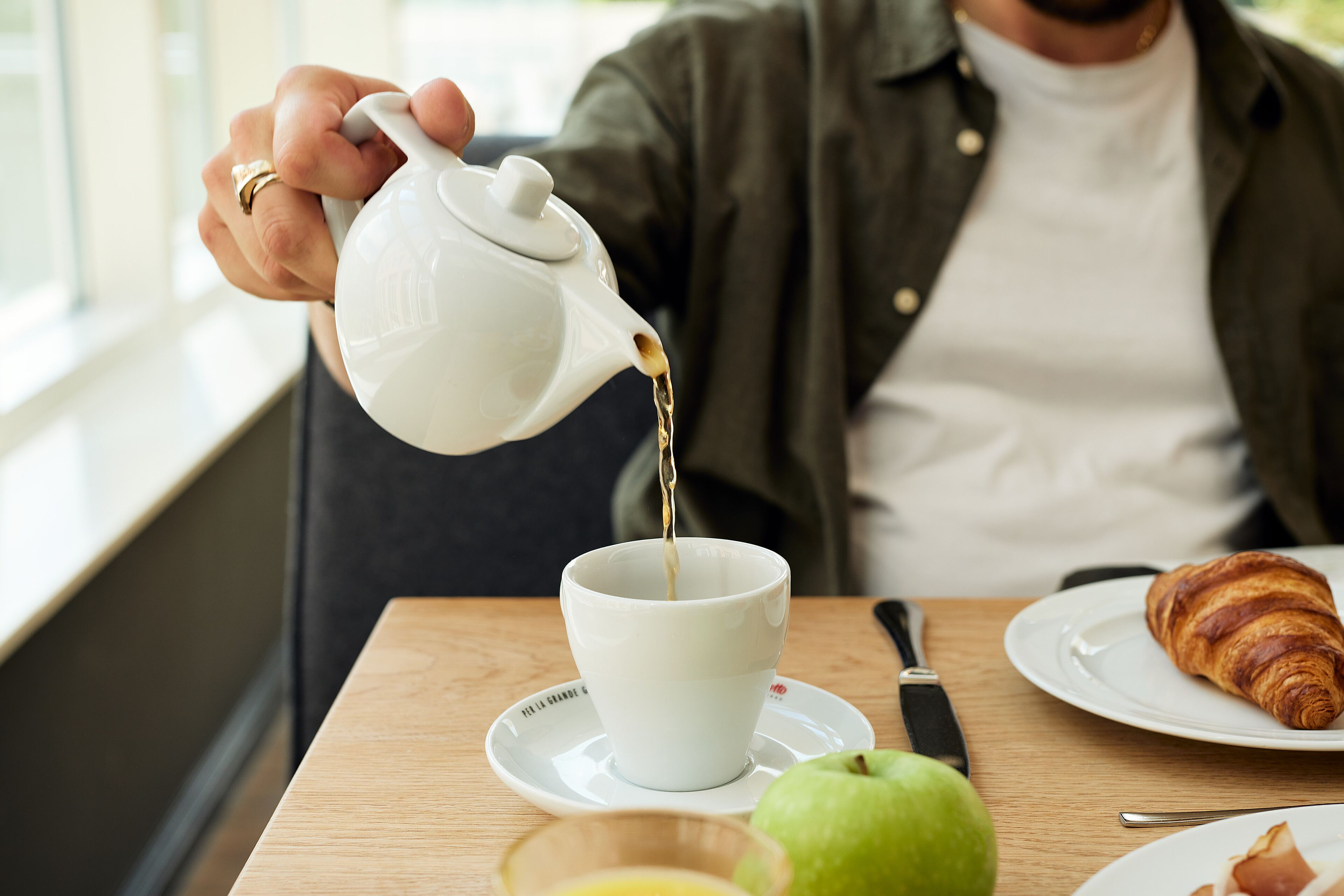 Una persona sirve té en una taza sobre una mesa con un croissant, una manzana y una naranja.