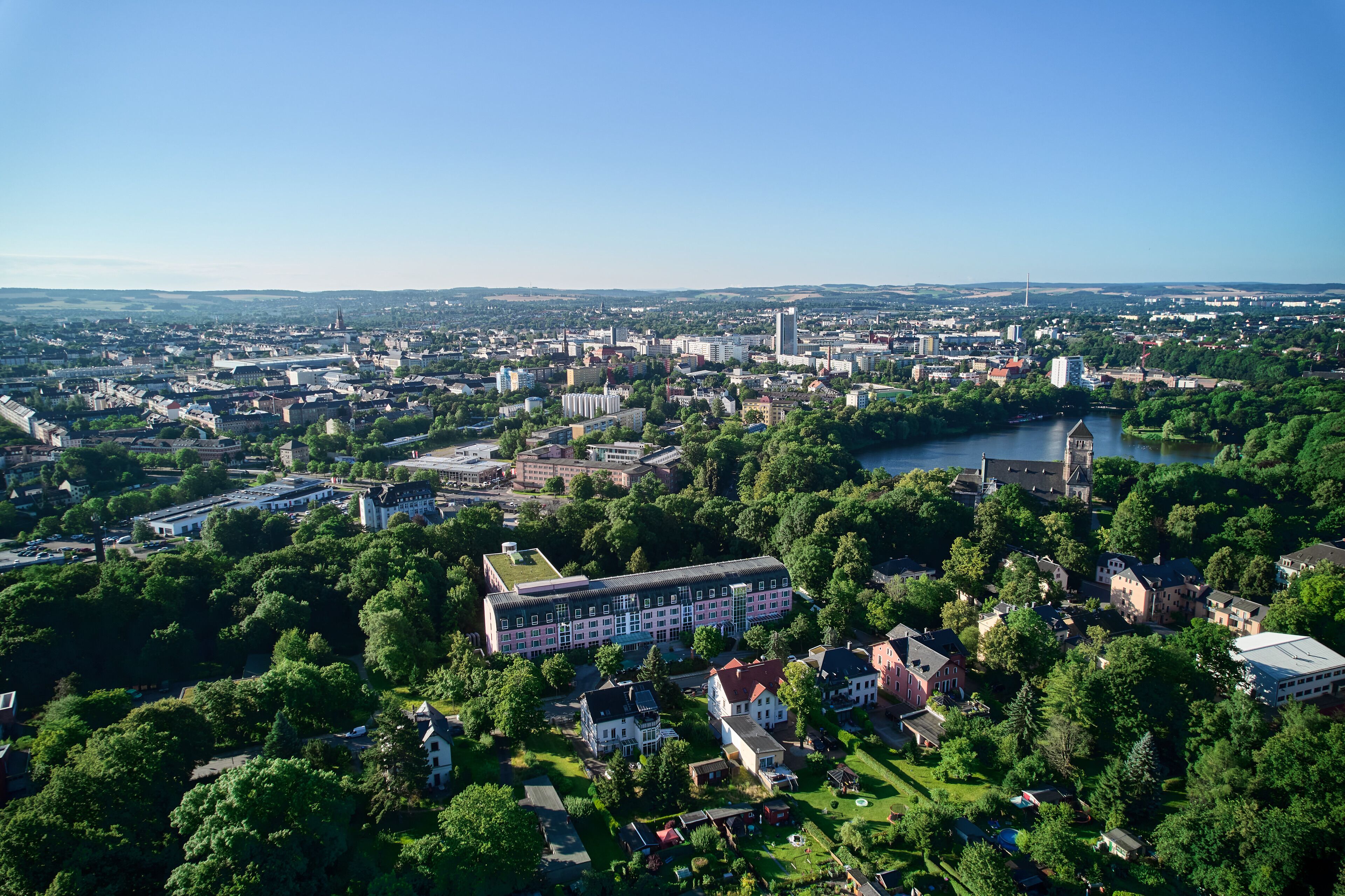 An aerial view of a city with a prominent building complex surrounded by greenery and a river.